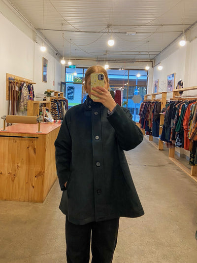 Person wearing a dark green coat in a clothing store with racks of clothes and a counter.