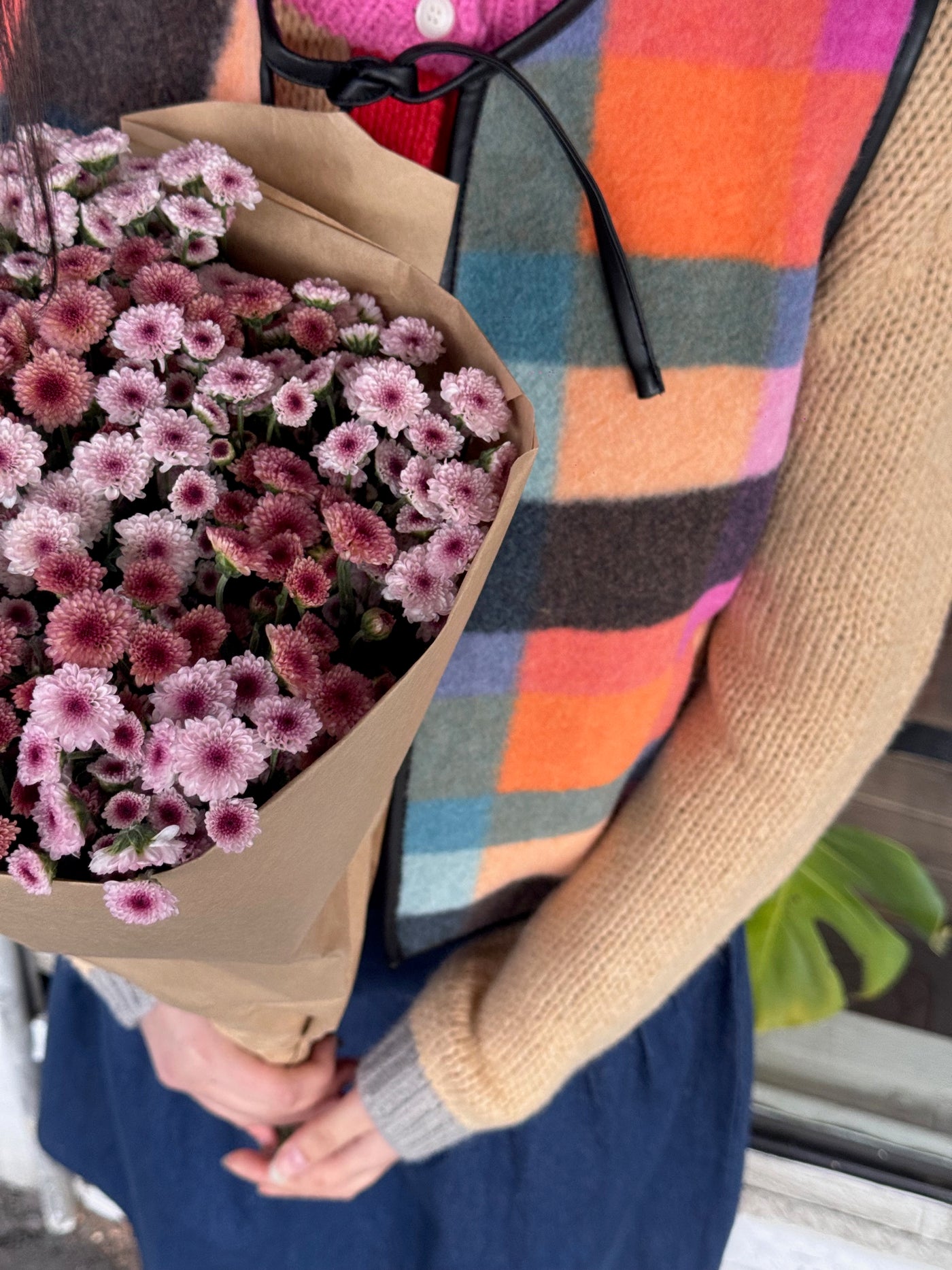 Person holding a bouquet of pink flowers wrapped in brown paper, wearing a colourful checked vest