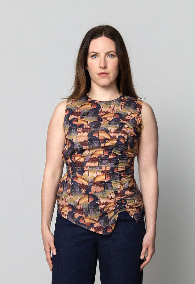 Woman wearing a sleeveless print top featuring a hand drawn toucan and dark blue flared denim pants on a plain background