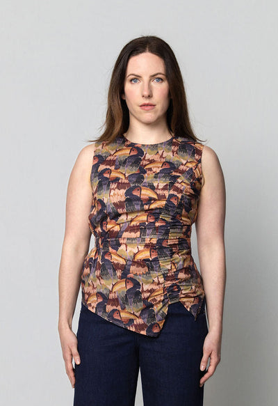 Woman wearing a sleeveless print top featuring a hand drawn toucan and dark blue flared denim pants on a plain background