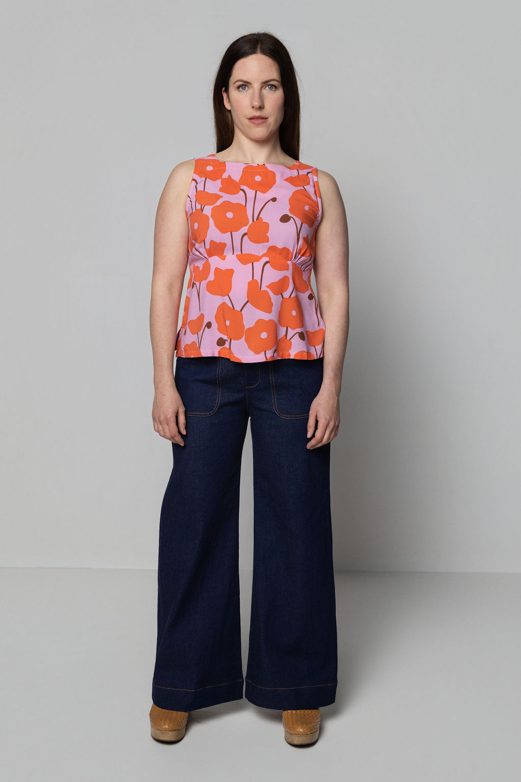 Woman wearing a sleeveless top with orange floral pattern and dark blue pants on a plain background