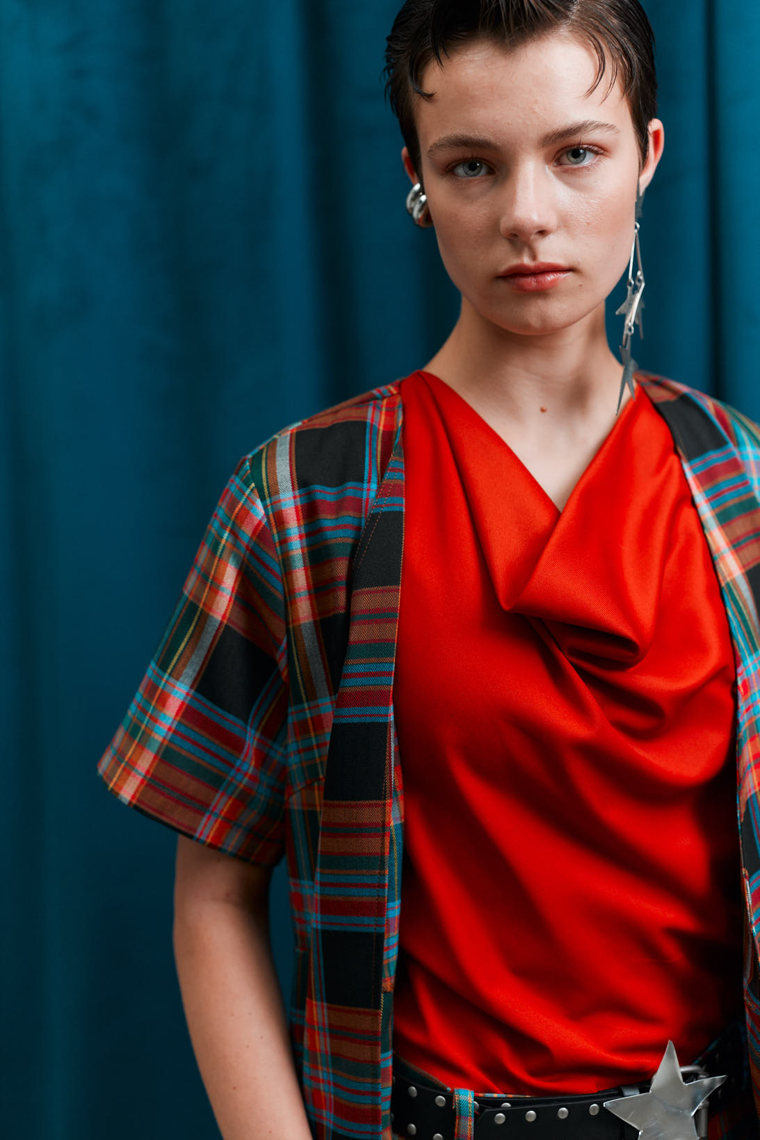 Person wearing a red top and plaid shirt against a blue curtain