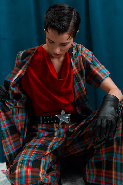 Person wearing a red top and plaid coat with matching pants and black gloves against a blue curtain.