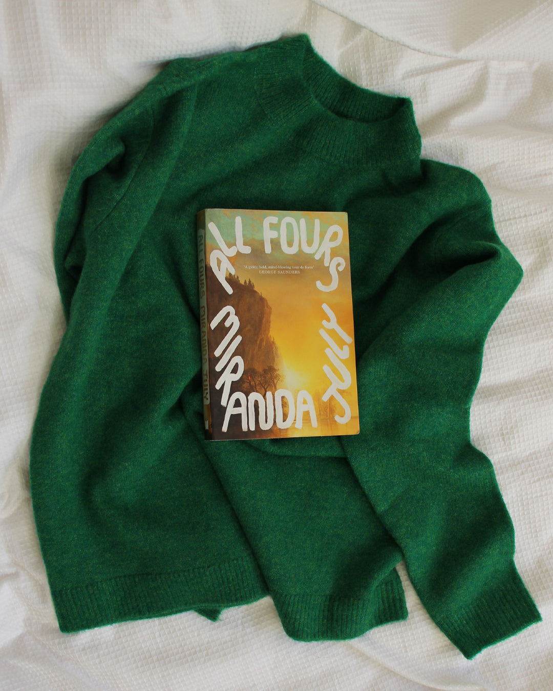 Green sweater with a book titled 'All Fours' by Miranda July on a white surface