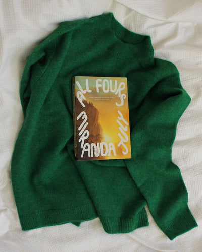 Green sweater with a book titled 'All Fours' by Miranda July on a white surface