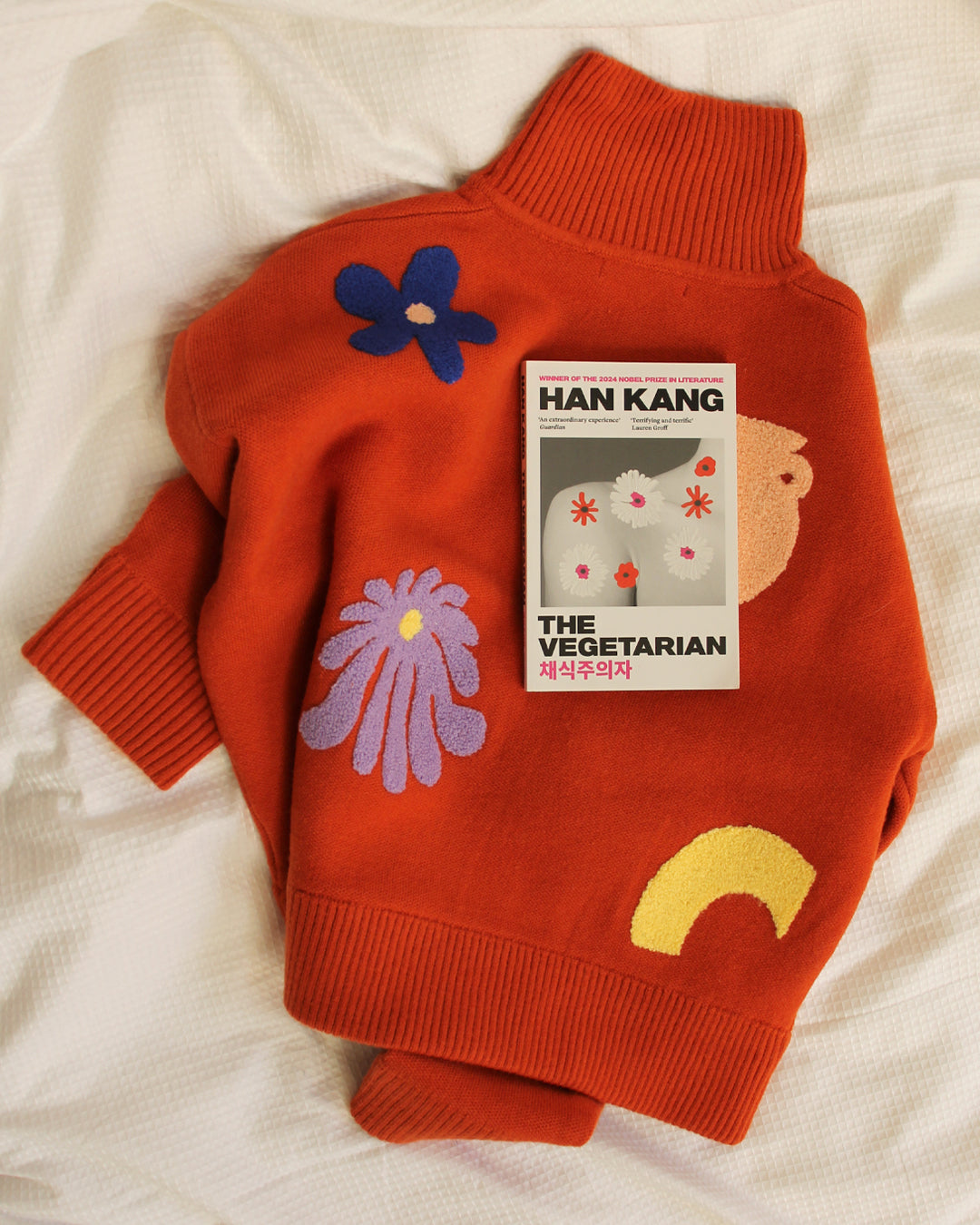 Orange sweater with floral designs and a book titled 'The Vegetarian' by Han Kang on a white background
