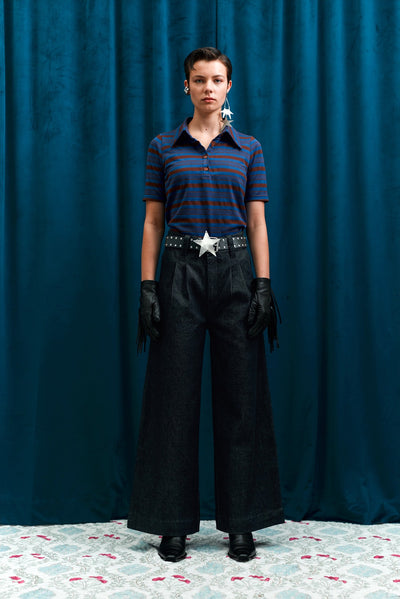 Person wearing a striped shirt and black pleated jeans with a belt and gloves, standing against a blue curtain.