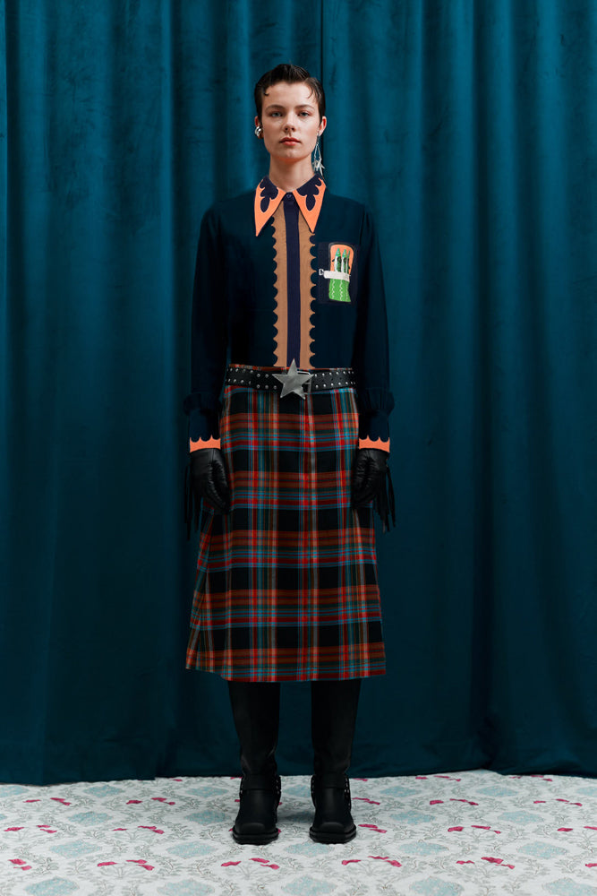 Person wearing a dark outfit with plaid skirt and orange accents against a blue curtain.