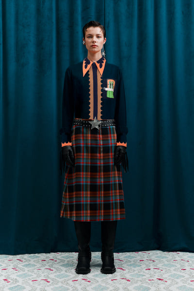 Person wearing a dark outfit with plaid skirt and orange accents against a blue curtain.