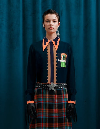 Person wearing a dark shirt with orange accents and a plaid skirt against a blue curtain.