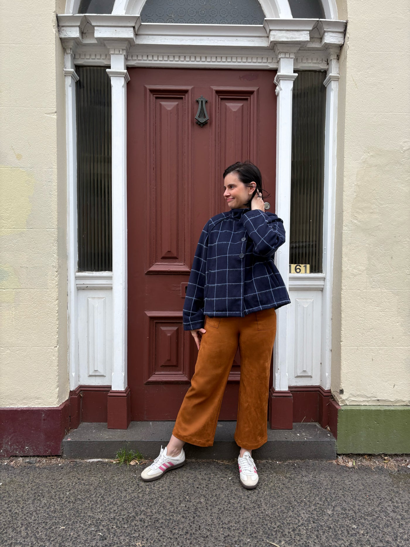 Person standing in front of a red door on a residential street wearing a navy checkered jacket and tan suede pants