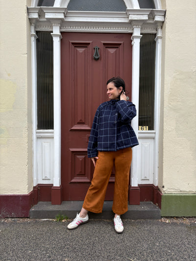 Person standing in front of a red door on a residential street wearing a navy checkered jacket and tan suede pants