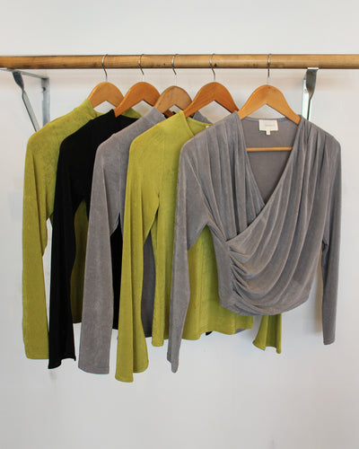 Four tops in green, black, and gray hanging on a wooden rack against a white wall.