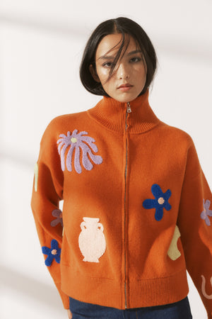 Person wearing A bright orange zip-up cardigan with tufted appliques of flowers and vases with blue jeans